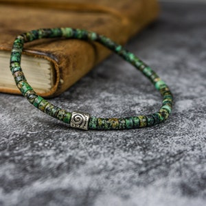 Puede incluir: Un brazalete de cuentas verdes y marrones con un dije de plata con un diseño en espiral.