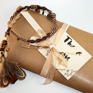 May include: A handcrafted macram&eacute; bracelet with feathers and beads, in shades of brown and beige, lies on a wrapped package. The package is tied with a ribbon and a tag that says "Thank you".