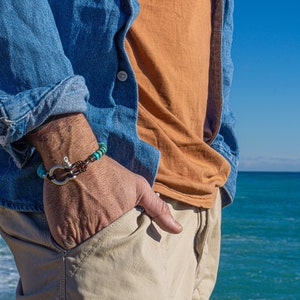 Op de afbeelding: Een man draagt een blauw denim shirt, een oranje t-shirt en een kaki broek. Hij draagt een turquoise en bruine kralen armband met een zilveren anker sluiting. De armband zit om zijn linker pols. Hij staat voor een blauwe zee.