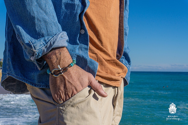 Op de afbeelding: Een man draagt een blauw denim shirt, een oranje t-shirt en een kaki broek. Hij draagt een turquoise en bruine kralen armband met een zilveren anker sluiting. De armband zit om zijn linker pols. Hij staat voor een blauwe zee.