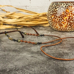 May include: Brown leather glasses chain with brown and gold beads. The chain is wrapped around a pair of brown sunglasses.