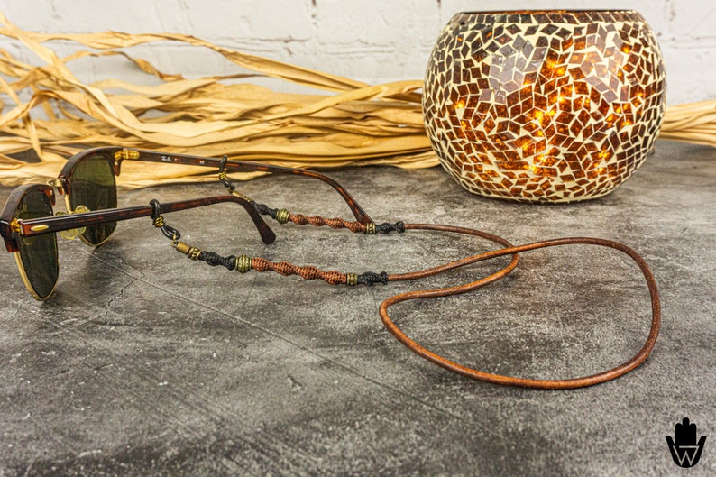 May include: Brown leather glasses chain with brown and gold beads. The chain is wrapped around a pair of brown sunglasses.