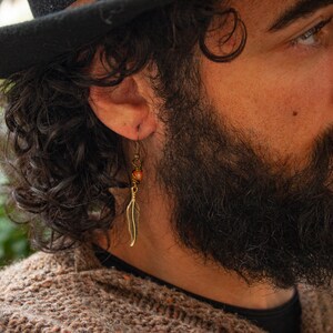 May include: A man with a black fedora and a long beard is wearing a gold feather earring with a brown bead.