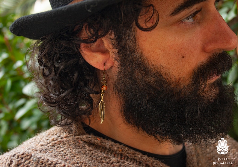 May include: A man with a black fedora and a long beard is wearing a gold feather earring with a brown bead.