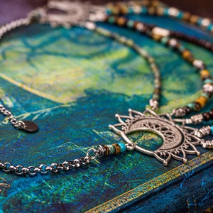 May include: A silver necklace with a crescent moon pendant and a beaded chain. The beads are turquoise, brown, and white. The necklace is lying on a blue and green surface.