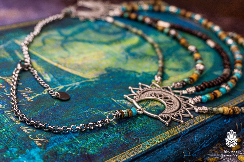 May include: A silver necklace with a crescent moon pendant and a beaded chain. The beads are turquoise, brown, and white. The necklace is lying on a blue and green surface.