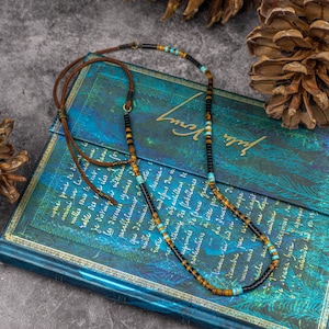 K&ouml;nnte beinhalten: Eine mehrfarbige Perlenkette mit braunen Lederkordel-Akzenten. Die Kette besteht aus schwarzen, braunen, t&uuml;rkisfarbenen und goldenen Perlen. Die Kette liegt auf einem blauen und gr&uuml;nen Buch mit goldenen Akzenten.