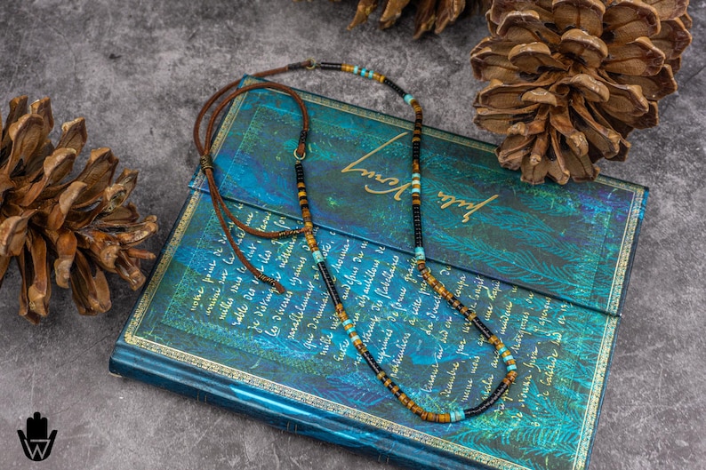 K&ouml;nnte beinhalten: Eine mehrfarbige Perlenkette mit braunen Lederkordel-Akzenten. Die Kette besteht aus schwarzen, braunen, t&uuml;rkisfarbenen und goldenen Perlen. Die Kette liegt auf einem blauen und gr&uuml;nen Buch mit goldenen Akzenten.