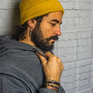 May include: A man wearing a gray hoodie with the hood up, a yellow beanie, and multiple bracelets on his wrist. He has a beard and is looking down.