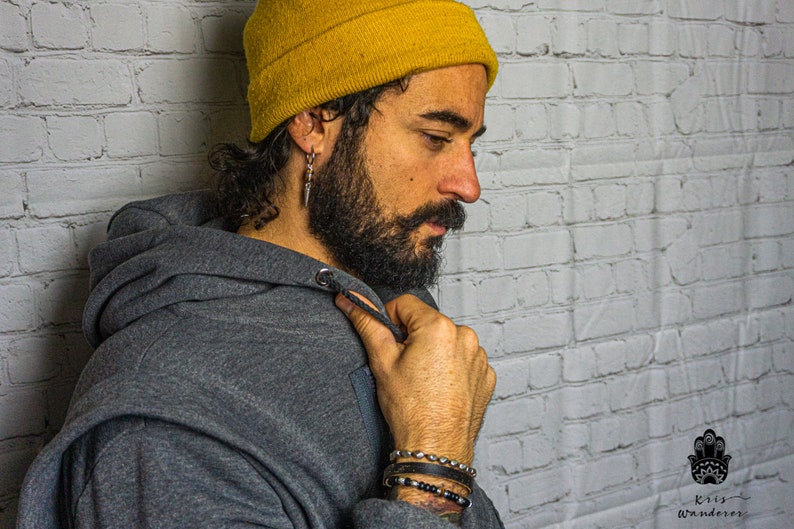 May include: A man wearing a gray hoodie with the hood up, a yellow beanie, and multiple bracelets on his wrist. He has a beard and is looking down.