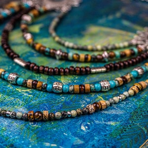 May include: A multi-strand necklace with brown wooden beads, turquoise beads, and silver accents. The necklace is on a blue and green patterned surface.