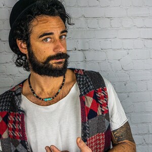 May include: A man with a black hat and a beard is wearing a multi-colored patchwork vest. He is wearing a necklace with black beads and a turquoise stone.