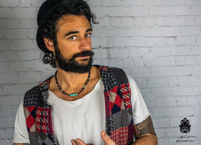 May include: A man with a black hat and a beard is wearing a multi-colored patchwork vest. He is wearing a necklace with black beads and a turquoise stone.