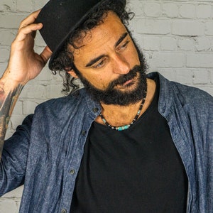 May include: A man with a beard and a black hat is wearing a blue denim shirt over a black t-shirt. He is wearing a beaded necklace with turquoise and black beads.