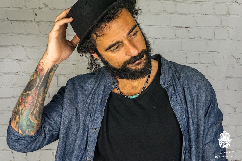 May include: A man with a beard and a black hat is wearing a blue denim shirt over a black t-shirt. He is wearing a beaded necklace with turquoise and black beads.