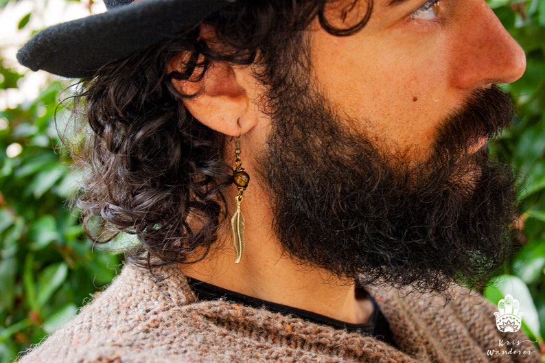 May include: A man with a beard and a black fedora is wearing a gold feather earring with a brown bead. The earring is hanging from his left ear.