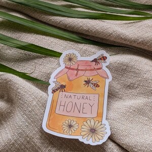 Peut inclure: Un autocollant repr&eacute;sentant un pot de miel avec un design aquarelle. Le pot est jaune et porte l'inscription "NATURAL HONEY". Des abeilles et des fleurs entourent le pot.