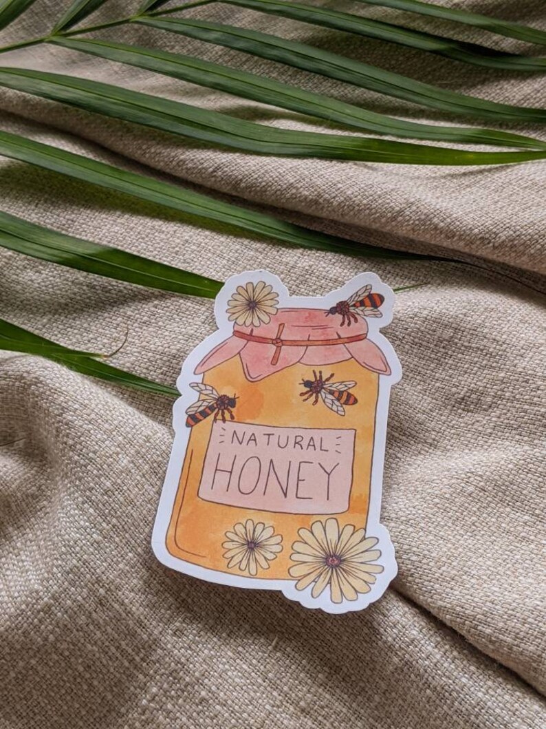 Peut inclure: Un autocollant repr&eacute;sentant un pot de miel avec un design aquarelle. Le pot est jaune et porte l'inscription "NATURAL HONEY". Des abeilles et des fleurs entourent le pot.