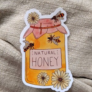Peut inclure: Un autocollant blanc avec une illustration de dessin anim&eacute; d'un pot de miel avec un couvercle rose. Le pot est rempli de miel jaune et porte une &eacute;tiquette blanche sur laquelle est &eacute;crit "NATURAL HONEY". Il y a trois abeilles et deux fleurs blanches autour du pot.