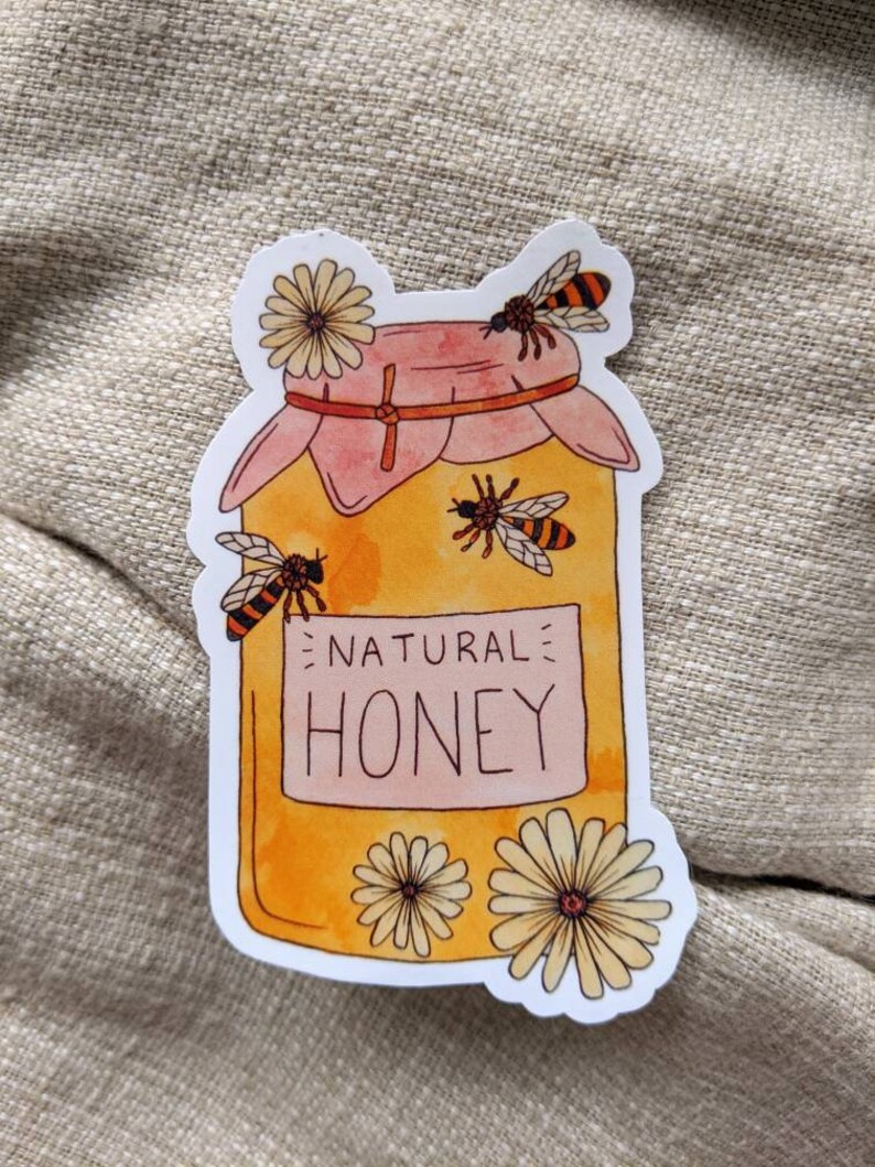 Peut inclure: Un autocollant blanc avec une illustration de dessin anim&eacute; d'un pot de miel avec un couvercle rose. Le pot est rempli de miel jaune et porte une &eacute;tiquette blanche sur laquelle est &eacute;crit "NATURAL HONEY". Il y a trois abeilles et deux fleurs blanches autour du pot.