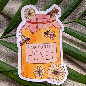 Peut inclure: Un autocollant repr&eacute;sentant un pot de miel avec un couvercle rose et une &eacute;tiquette jaune sur laquelle est inscrit "NATURAL HONEY". Le pot est entour&eacute; d'abeilles et de fleurs.