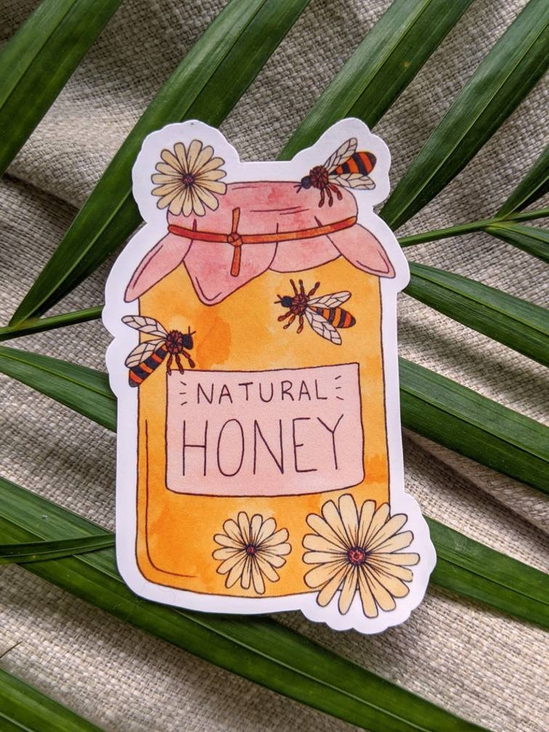 Peut inclure: Un autocollant repr&eacute;sentant un pot de miel avec un couvercle rose et une &eacute;tiquette jaune sur laquelle est inscrit "NATURAL HONEY". Le pot est entour&eacute; d'abeilles et de fleurs.