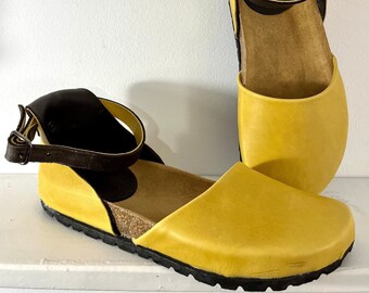 May include: A pair of yellow leather sandals with brown leather straps and a cork footbed. The sandals have a black rubber sole.