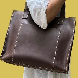 May include: A large brown leather tote bag with a front pocket and white stitching. The bag has a top handle and two shoulder straps.