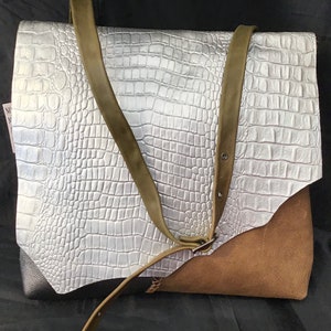 Puede incluir: Un bolso tote de cuero plateado y marrón con un patrón de cocodrilo en relieve. El bolso tiene una correa larga y un cierre de solapa.