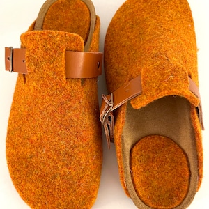 Könnte beinhalten: Ein Paar orangefarbene Filzclogs mit braunen Lederriemen und Schnallen. Die Clogs haben ein strukturiertes Aussehen und ein konturiertes Fußbett. Die Riemen sind verstellbar. Die Clogs scheinen ungetragen zu sein.