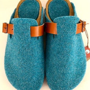 May include: A pair of teal blue felt slippers with brown leather straps and buckles. The slippers have a rounded toe and a comfortable, casual look.