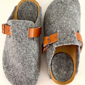 Può includere: Un paio di zoccoli in feltro grigio con cinturini e fibbie in pelle marrone. Gli zoccoli hanno una punta arrotondata e un tacco leggermente rialzato.