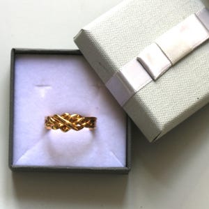 May include: A gold ring with an interwoven design, displayed in a grey gift box with a white ribbon. The ring's detailed pattern is set against the box's light interior, suggesting a jewellery item.