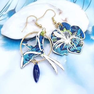 May include: A pair of dangle earrings featuring a blue and white floral design and a blue and white bird design with a gold ring and a blue teardrop charm.
