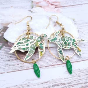 Peut inclure: Boucles d'oreilles créoles dorées avec un pendentif tortue à motif vert et blanc et un pendentif feuille verte.