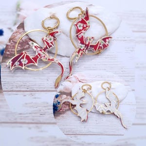Könnte beinhalten: Drachenförmige Ohrringe mit rotem Blumenmuster und goldfarbenen Reifen. Die Flügel des Drachen sind ausgebreitet, und das Design ist mit weißen und goldenen Akzenten versehen. Die Ohrringe werden auf einer weißen Oberfläche präsentiert.