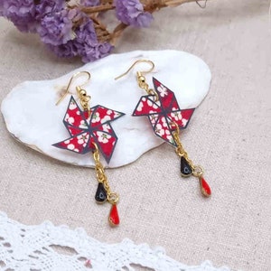 May include: Pinwheel-shaped earrings with a red floral pattern and gold-colored hooks. Each earring has a black and red teardrop charm hanging from a gold chain. The earrings are displayed on a white, shell-shaped surface.