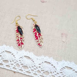 Puede incluir: Un par de pendientes de oro con colgantes en forma de plumas rojas y blancas y cuentas negras.