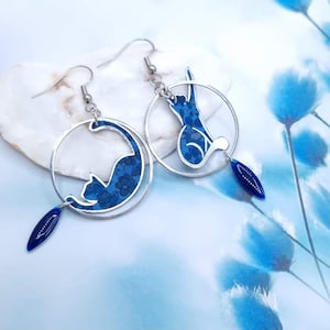 May include: A pair of silver hoop earrings with a blue cat silhouette inside each hoop. Each earring has a small blue teardrop charm hanging from the bottom.