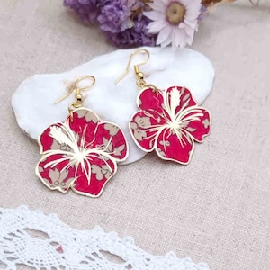 Peut inclure: Une paire de boucles d'oreilles en or avec des motifs de fleurs d'hibiscus rouges. Les fleurs ont un motif blanc et or.