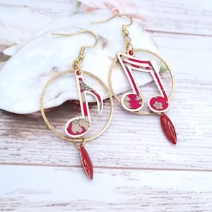 May include: Gold-coloured hoop earrings with musical note pendants. The notes are red with a floral pattern and white trim, with a red teardrop-shaped charm dangling below. The earrings are displayed on a white wooden surface.