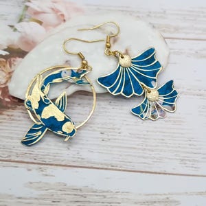 Puede incluir: Un par de pendientes de oro con esmalte azul y blanco. Un pendiente presenta un diseño de pez koi estilizado, mientras que el otro presenta un par de hojas de ginkgo.