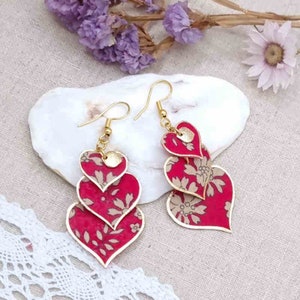 May include: A pair of gold tone earrings with three layered red and gold heart-shaped charms. The hearts have a floral pattern.