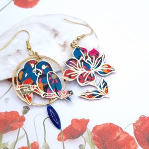 Pode incluir: Um par de brincos coloridos com um design floral e dois papagaios. Os brincos são feitos de papel e têm um acabamento dourado.