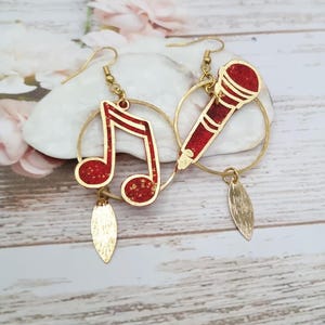 Peut inclure: Une paire de boucles d'oreilles créoles dorées avec des accents rouges et dorés. Une boucle d'oreille présente un charme de note de musique et l'autre un charme de microphone. Chaque boucle d'oreille a une petite feuille d'or pendante.