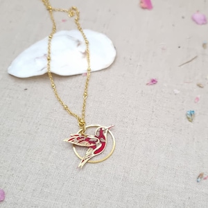 May include: A gold chain necklace with a red and gold hummingbird pendant. The hummingbird is in flight and is surrounded by a gold circle.