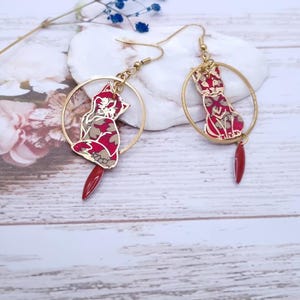 May include: A pair of gold-tone cat earrings. Each earring features a red and patterned cat silhouette within a gold hoop, with a dangling red teardrop. The earrings are set against a soft, floral background.