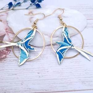 Può includere: Un paio di orecchini a cerchio color oro con un design a uccello blu e bianco. Gli uccelli hanno un motivo dettagliato e sono attaccati ai cerchi con ganci color oro. Gli orecchini sono esposti su una superficie bianca.