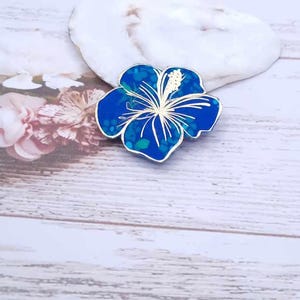 Pode incluir: Um broche azul em forma de flor de hibisco com contorno dourado e detalhes brancos. O broche tem uma aparência texturizada e salpicada no centro. O fundo inclui uma pedra branca e uma superfície de madeira.
