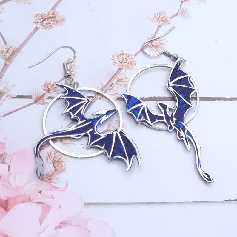 May include: A pair of silver hoop earrings with a blue dragon design. The dragon is flying in a circle and has a detailed, textured finish.